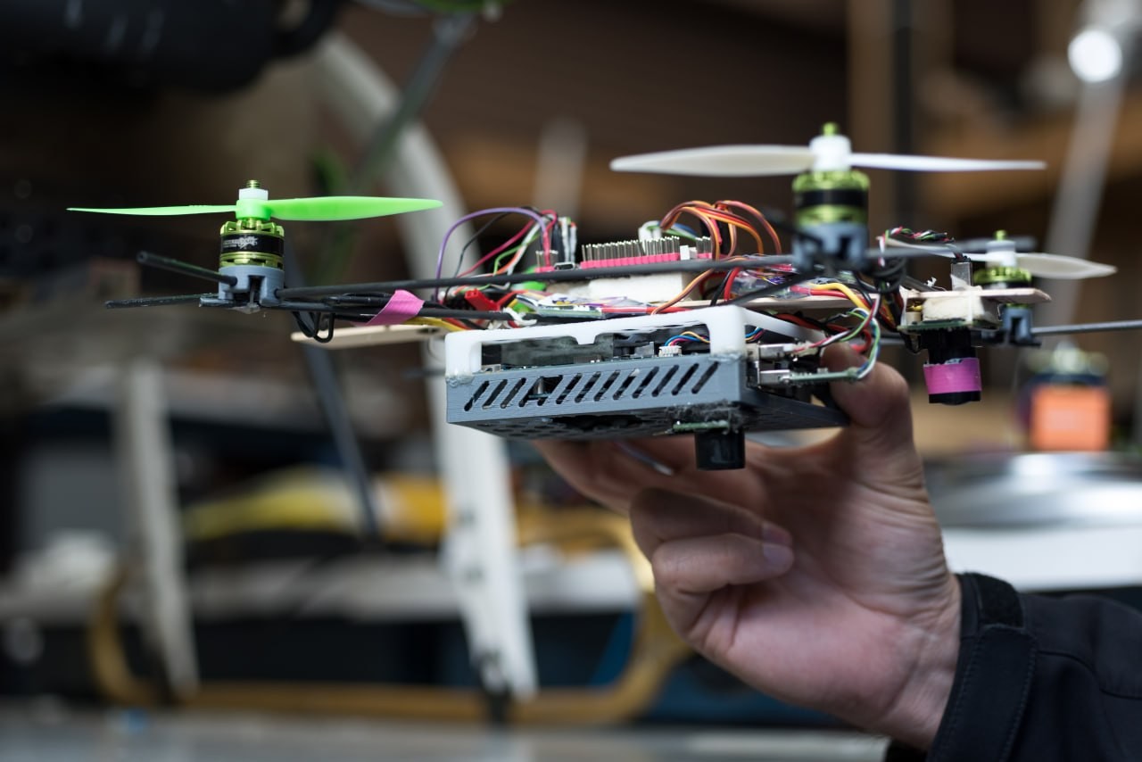 A hand holds a prototype drone with exposed electronics and wiring.